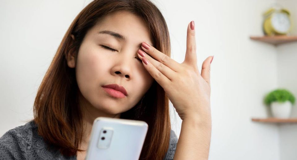 Woman holding her smartphone and rubbing her eyes, showing signs of eye strain and fatigue from screen use.